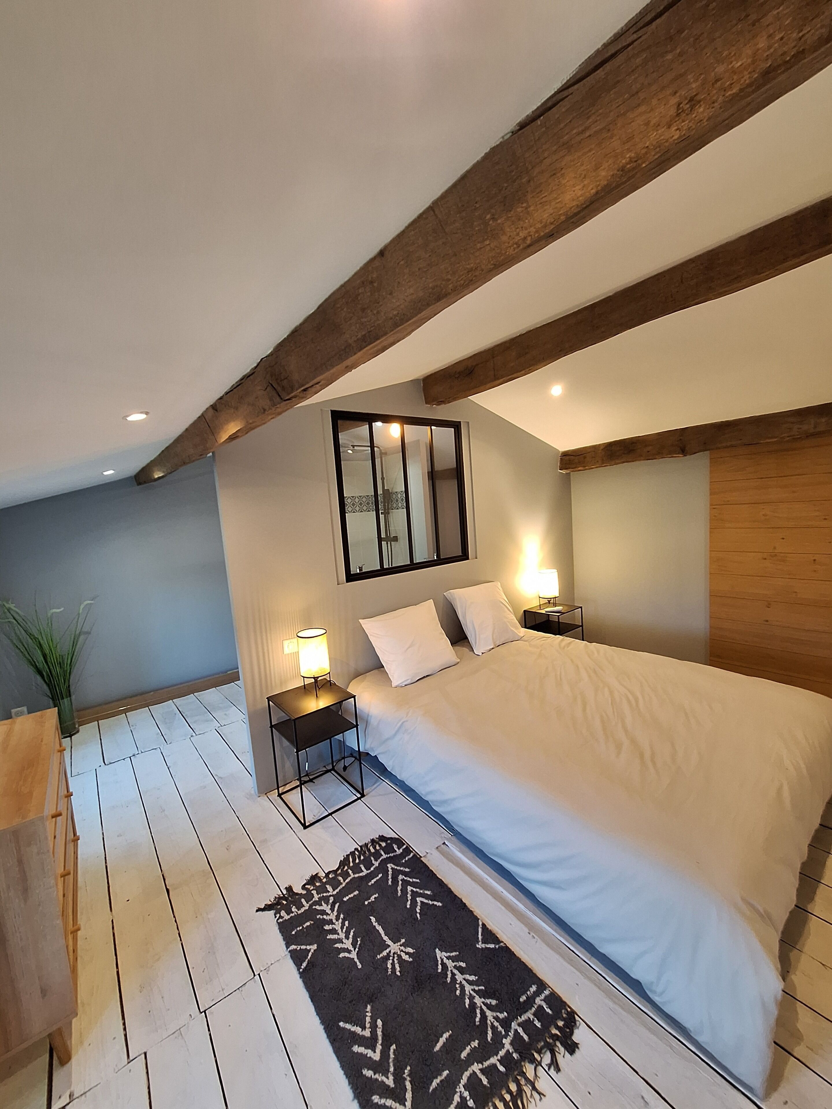 A bedroom with a bed, bedside tables, a nightstand, a mirror, a wooden ceiling beam, and a potted plant.