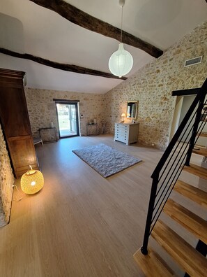 A spacious room with a wooden floor, a stone wall, a wooden cabinet, a mirror, a hanging light fixture, and a staircase.