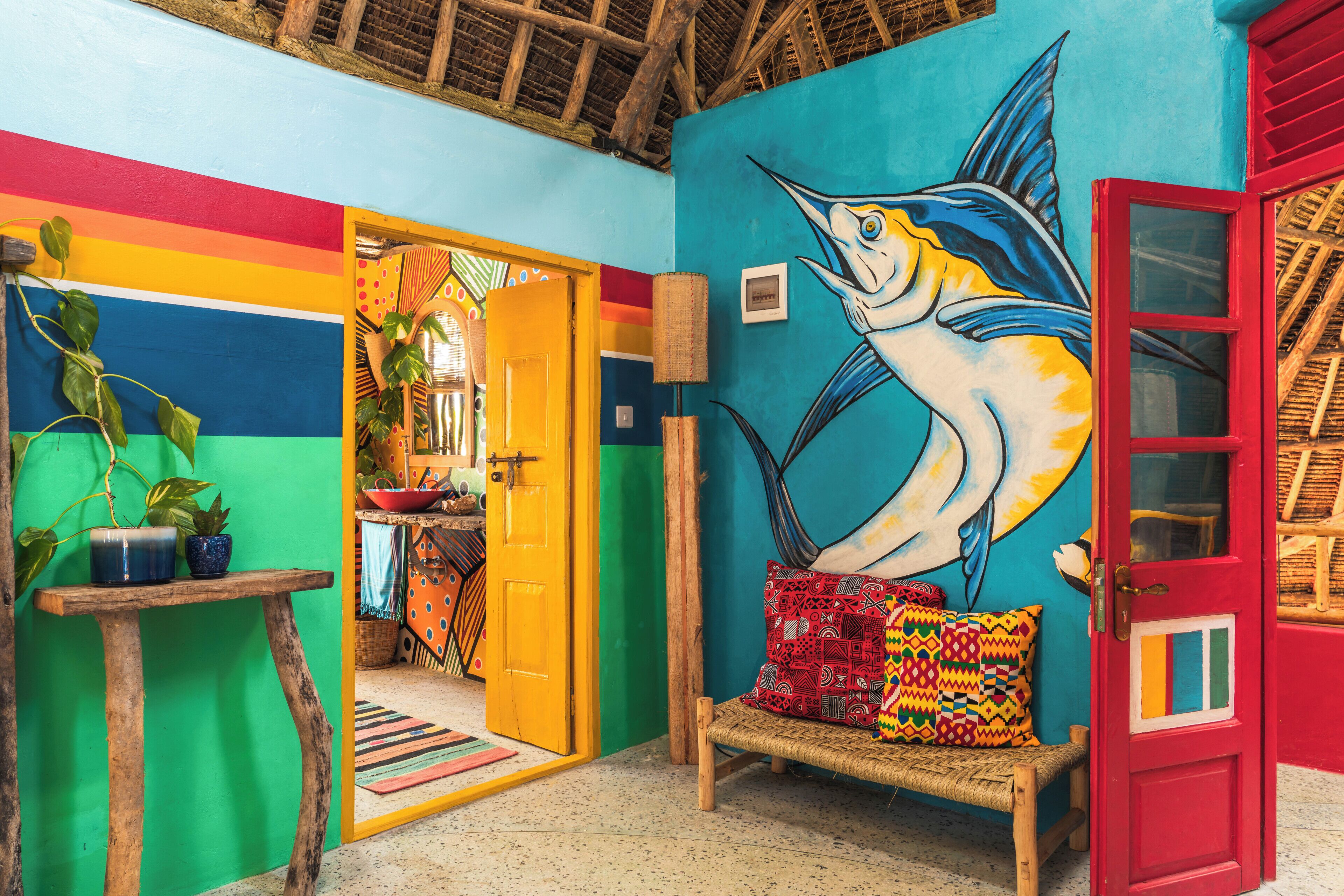 Una habitación vibrante con un mural de pared colorido, un banco de madera con cojines estampados, una puerta roja y un banco tejido.
