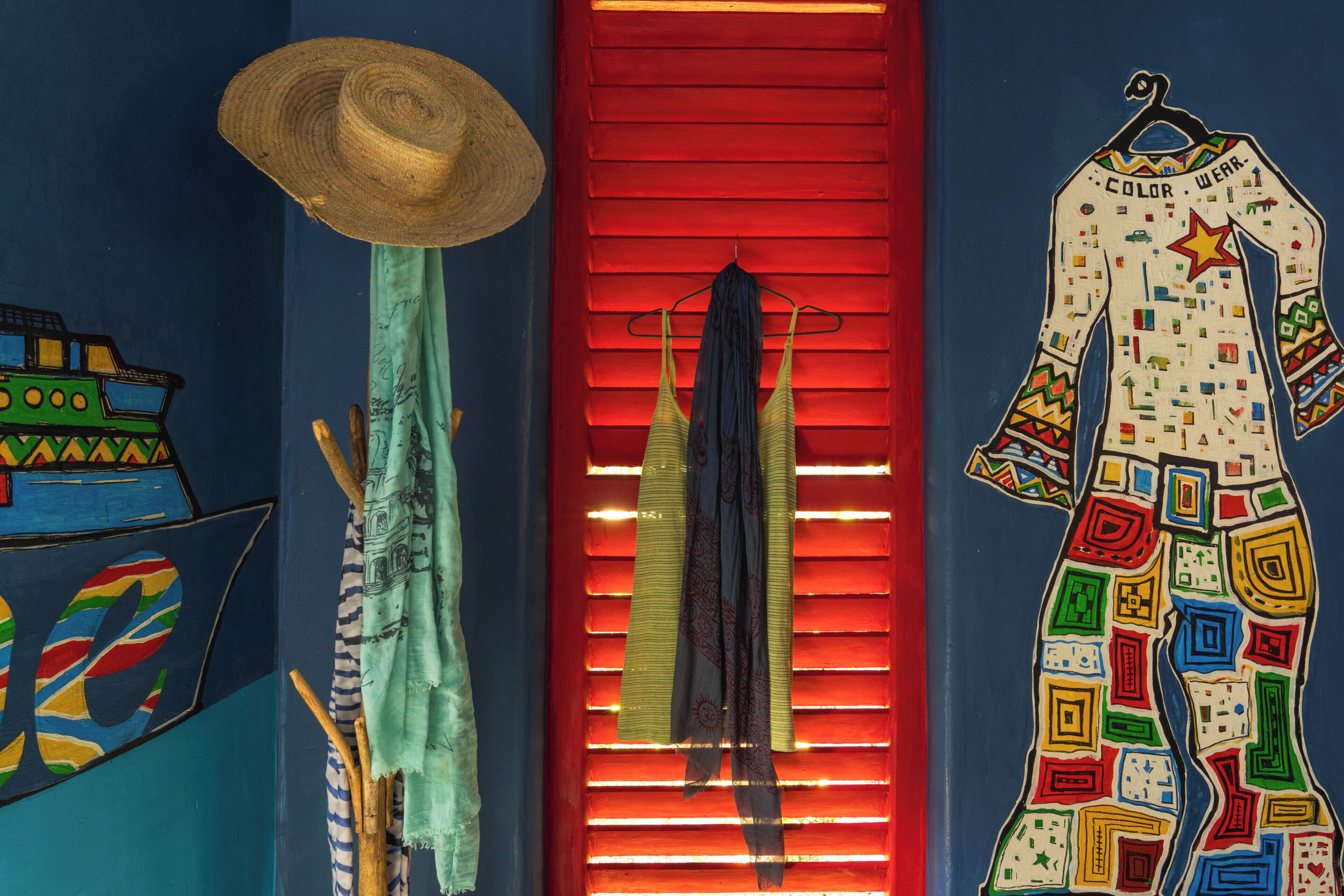 Una habitación con un persiano rojo, un sombrero, un vestido con estampados coloridos y una pared con un mural multicolor.
