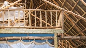 Una habitación con techo de paja, escalera de madera, banco acolchado azul y cama con dosel blanco.