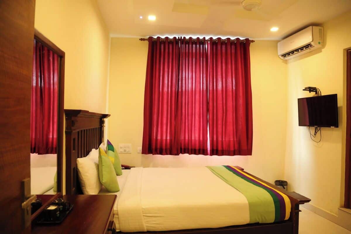 A hotel room with a bed, a TV, an air conditioning unit, and a window with red curtains.