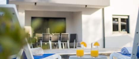 A poolside area with lounge chairs, a table with two glasses of orange juice, and a modern building in the background.