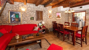 Una sala de estar acogedora con un sofá rojo, una mesa de comedor de madera, paredes de piedra y una cocina visible a través de un vano abierto.
