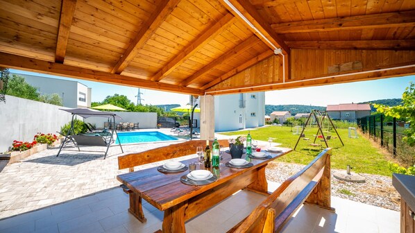 An outdoor dining area with a wooden table and benches, a pool, and a view of a residential area.