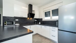 A modern kitchen with white cabinets, a black countertop, a stainless steel refrigerator, and a window with blinds.