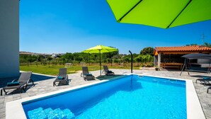 En swimmingpool med liggestole og en grøn parasol, omgivet af græs og en bygning med et tagtækket tag.