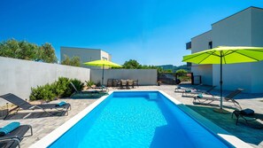 A rectangular swimming pool with lounge chairs and umbrellas, surrounded by a low wall and vegetation.