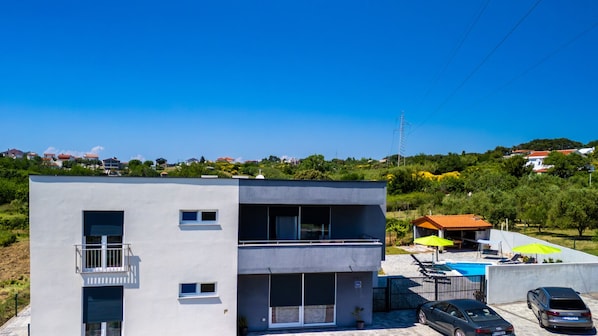 Un edificio moderno de dos pisos con balcón, un área de piscina con sillas de descanso y sombrillas, y un estacionamiento con automóviles.