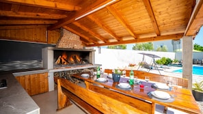 Una cocina al aire libre con horno de ladrillo, mesa de madera dispuesta para comer y una piscina al fondo.