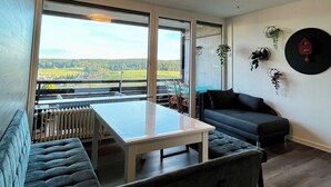 A modern living room with a large window, a sofa, a table, and a wall-mounted clock.