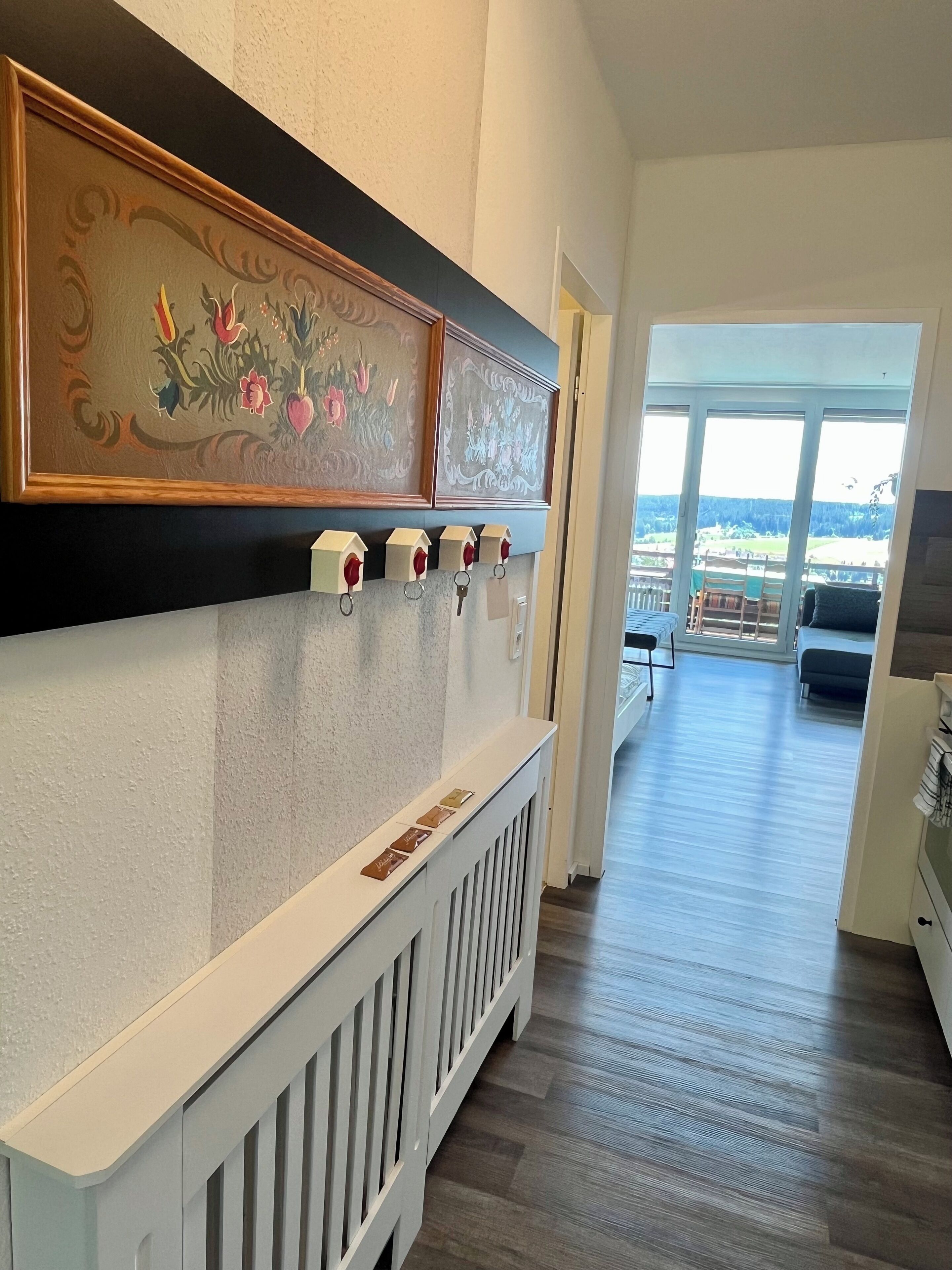 Ein Flur mit einem weißen Heizkörper, einem Kunstwerk mit Holzrahmen und Blick auf einen Balkon mit Gartenmöbeln.