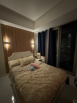 A bedroom with a bed, a nightstand, a window with curtains, and a view of a building outside. - The Bawhaus (Parañaque)