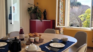 Un coin repas avec une table dressée pour quatre personnes, une fenêtre donnant sur un bâtiment et des arbres, et un meuble rouge orné d’une plante.