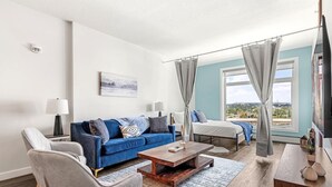 Ein modernes Wohnzimmer mit einem blauen Sofa, einem Holztisch und einem grauen Sessel. Ein Bett mit grauen Vorhängen und Blick ins Freie.