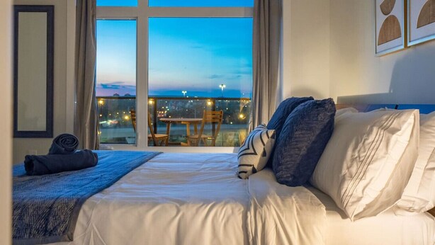 Ein Hotelzimmer mit einem großen Bett, Blick auf die Stadt im Abendlicht und einem Balkon mit Tisch und Stühlen.