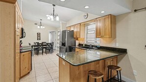 A kitchen with wooden cabinets, a granite countertop, a refrigerator, and a dining area with chairs.