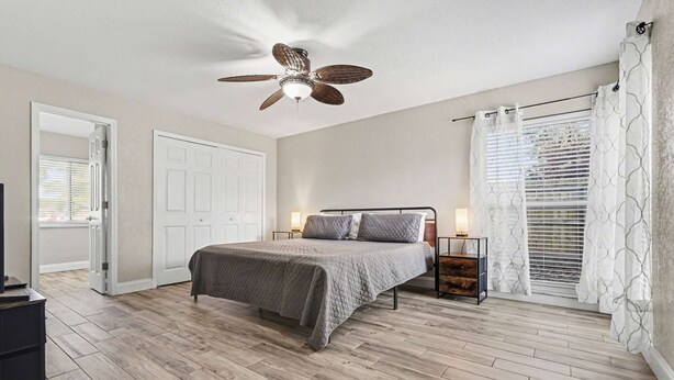 A bedroom with a bed, a ceiling fan, a bedside table with a lamp, a window with blinds, and a door.