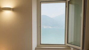 A hotel room with a bed, two towels, a wooden table, a wall-mounted light, and a window with a view of a body of water and mountains. - Lake front house with lake view and garage (Pisogne)