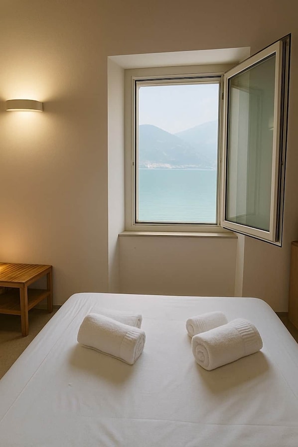 A hotel room with a bed, two towels, a wooden table, a wall-mounted light, and a window with a view of a body of water and mountains.