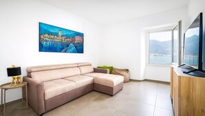 A living room with a beige sectional sofa, a small side table with a lamp, a painting of a cityscape, a large window with a view, and a wooden cabinet. - Lake front house with lake view and garage (Pisogne)