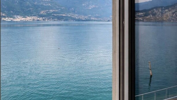 A view through a window of a calm lake with mountains in the background. - Lake front house with lake view and garage (Pisogne)