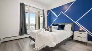 A modern bedroom with a large bed, a nightstand, a dresser, and a view of the outdoors through a window. - Lux Downtown Urban Escape W/ Gym & 2 Free Parking (Calgary)
