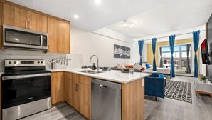 A modern kitchen with a dishwasher, oven, and microwave. There is a dining area with a sofa and a view of the outdoors. - Lux Downtown Urban Escape W/ Gym & 2 Free Parking (Calgary)