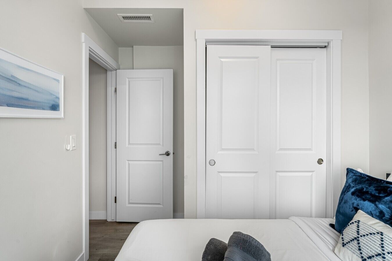 A bedroom with a bed, two white doors, a hallway, and a painting on the wall.