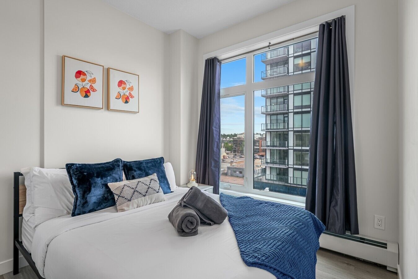 Un dormitorio con cama, dos obras de arte en la pared, una ventana con cortinas y vistas a edificios.