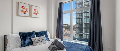Un dormitorio con cama, dos obras de arte en la pared, una ventana con cortinas y vistas a edificios.