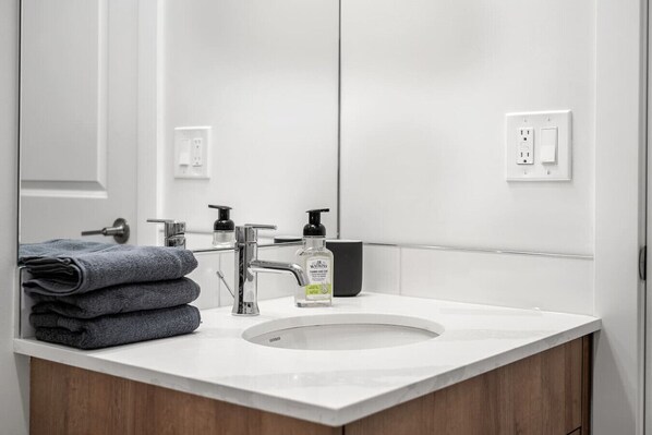 A bathroom with a white sink, a mirror, a towel stack, and a bottle of hand soap.