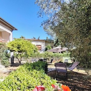 A garden with a patio area, a tree, and a building in the background. - Vacation house Audrey, three-room apartment in detached house with garden (Porto Azzurro)
