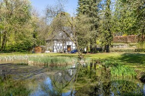 Ein Haus mit Teich davor, umgeben von Bäumen und einem klaren Himmel.