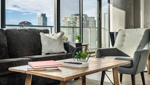En moderne stue med sofa, en grå lænestol, et træsofabord og udsigt til byens skyline gennem store vinduer.