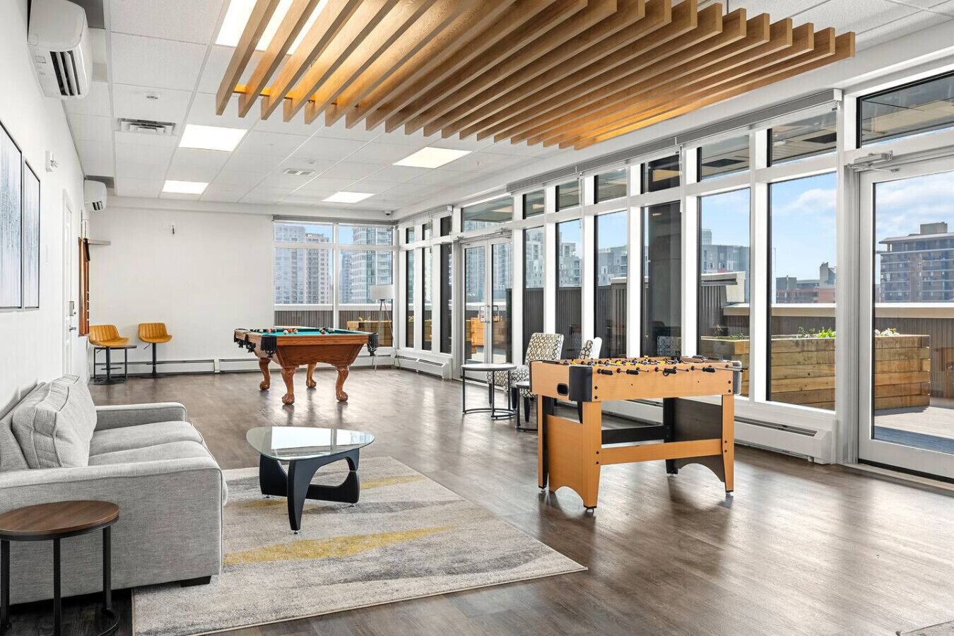 A spacious room with a pool table, foosball table, and a sofa. Large windows offer views of the city.