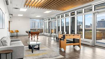 A spacious room with a pool table, foosball table, and a sofa. Large windows offer views of the city.