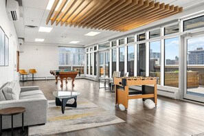 A spacious room with a pool table, foosball table, and a sofa. Large windows offer views of the city.