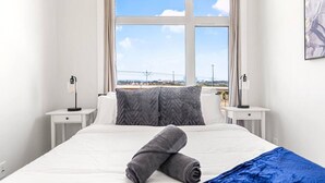 A hotel room with a bed, two bedside tables, a window with a view, and a painting on the wall. - Luxe Downtown Haven W / 2 Free Parking! (Calgary)