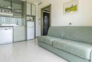 A compact kitchen with a dishwasher, a refrigerator, and a sink. There is a green sofa with floral patterns and a framed picture on the wall. - Residence Hotel Club House (Cattolica)