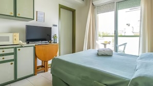 A hotel room with a bed, a desk with a TV, a small table with a plant, and a balcony with a chair. - Residence Hotel Club House (Cattolica)