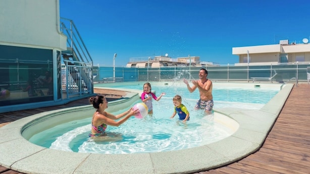 A swimming pool area with a wooden deck, a glass enclosure, and a clear blue sky. - Residence Hotel Club House (Cattolica)
