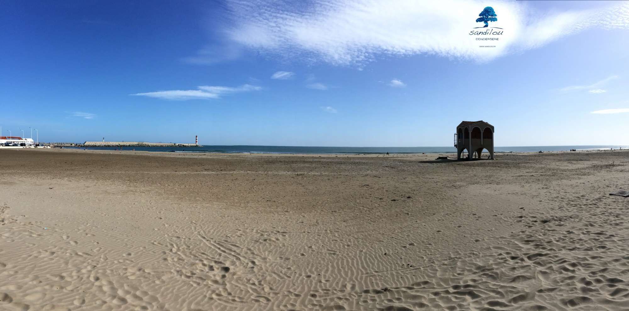 Una playa ancha y arenosa con huellas, un faro y una torre de salvavidas a lo lejos.