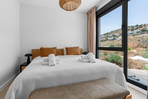 Ein Schlafzimmer mit einem großen Bett, Blick auf eine hügelige Landschaft und einem Fenster mit Vorhängen.
