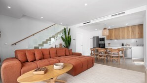 A modern living room with a sectional sofa, a dining area with wooden chairs, a kitchen with white cabinets, and a staircase with glass railings. - 7 - Bright house with private plunge pool (Mijas)