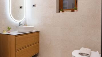A modern bathroom with a wooden vanity, a white sink, a mirror with an oval frame, a small window, and a white toilet.
