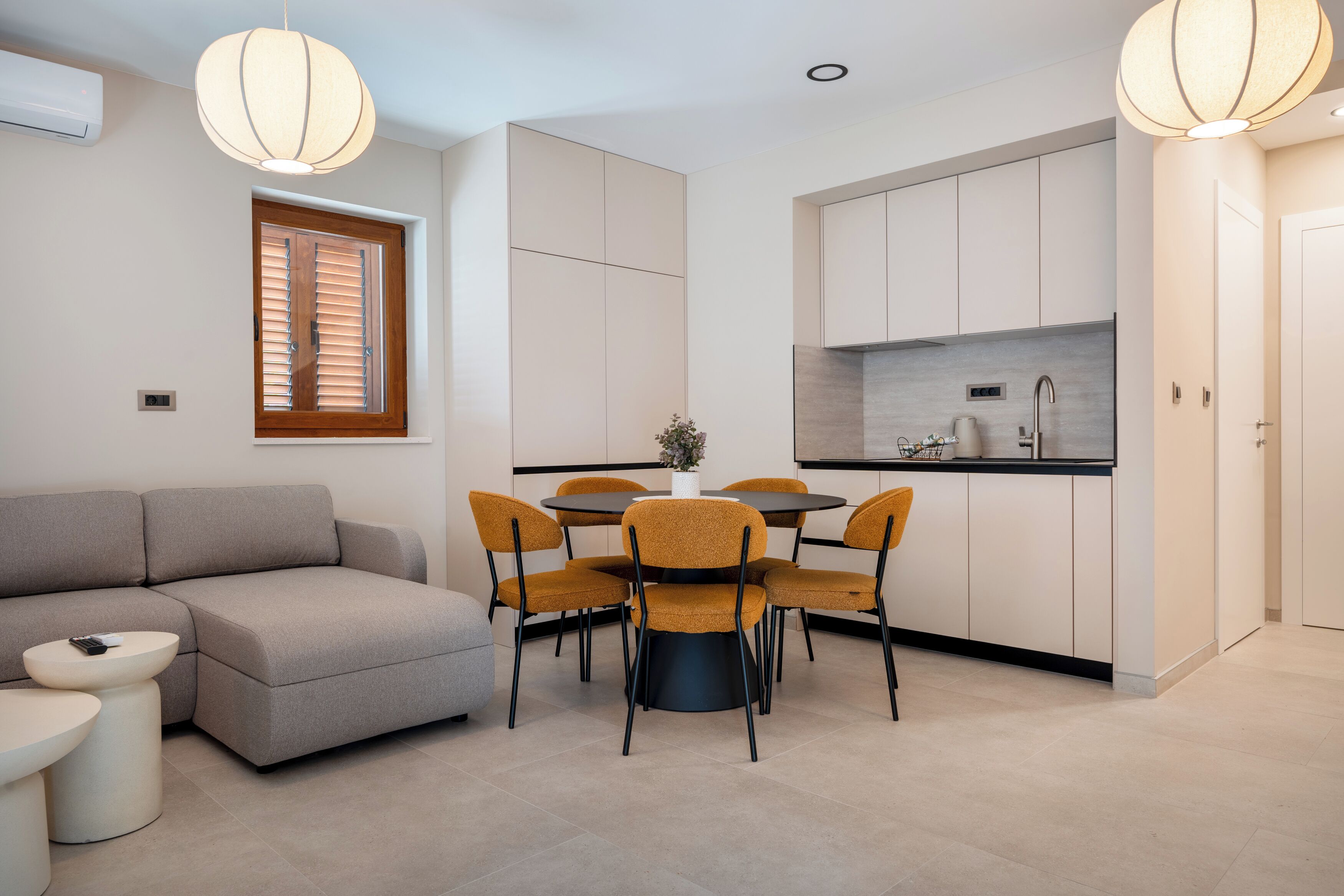 A modern living space with a sofa, round dining table, and chairs, a kitchen area with cabinets, and pendant lighting.