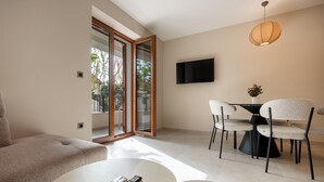 A modern living room with a sofa, a dining table with chairs, a TV mounted on the wall, and a sliding glass door leading to an outdoor area.