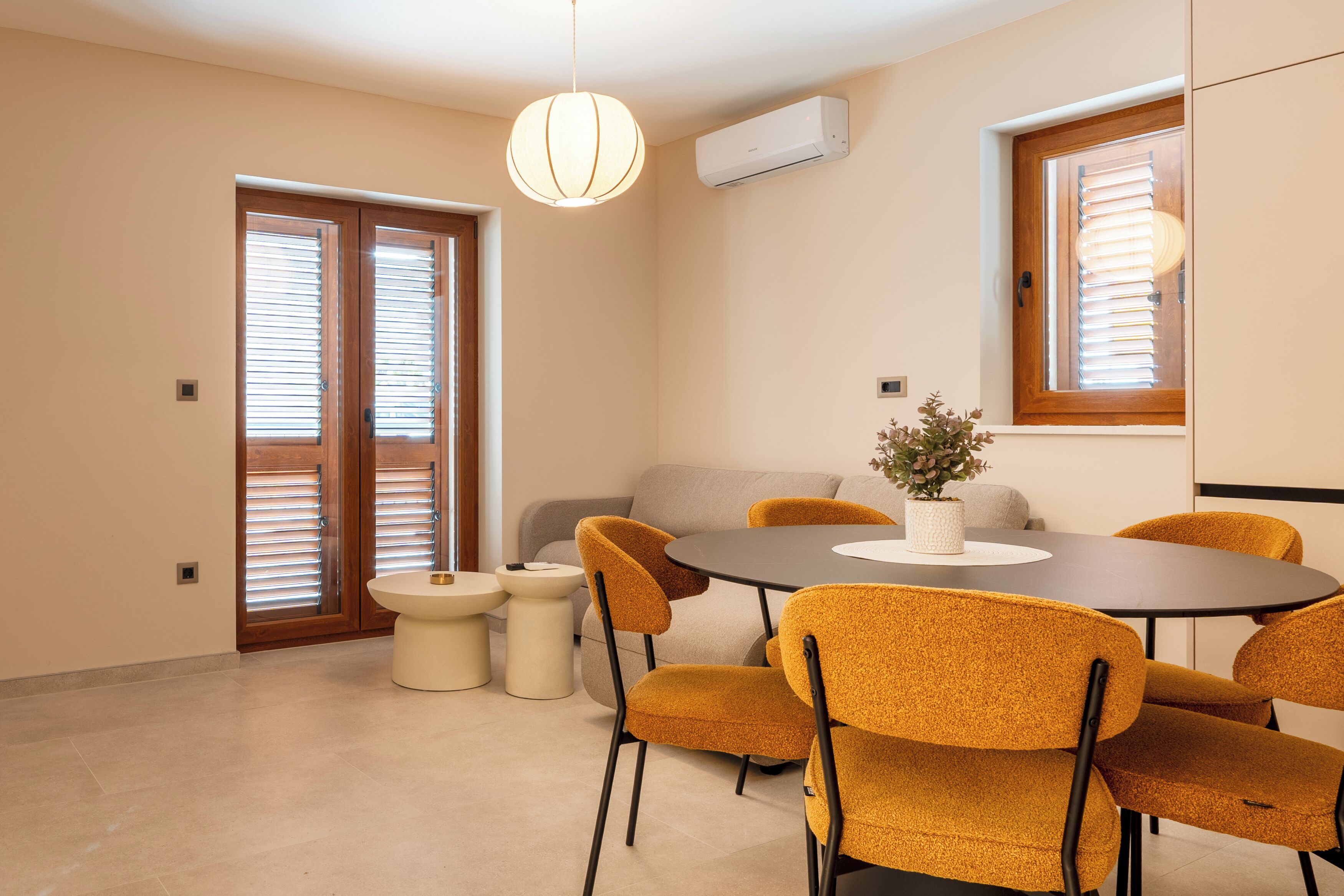 Uma sala de jantar com uma mesa redonda, seis cadeiras, uma mesinha lateral e um vaso com planta.