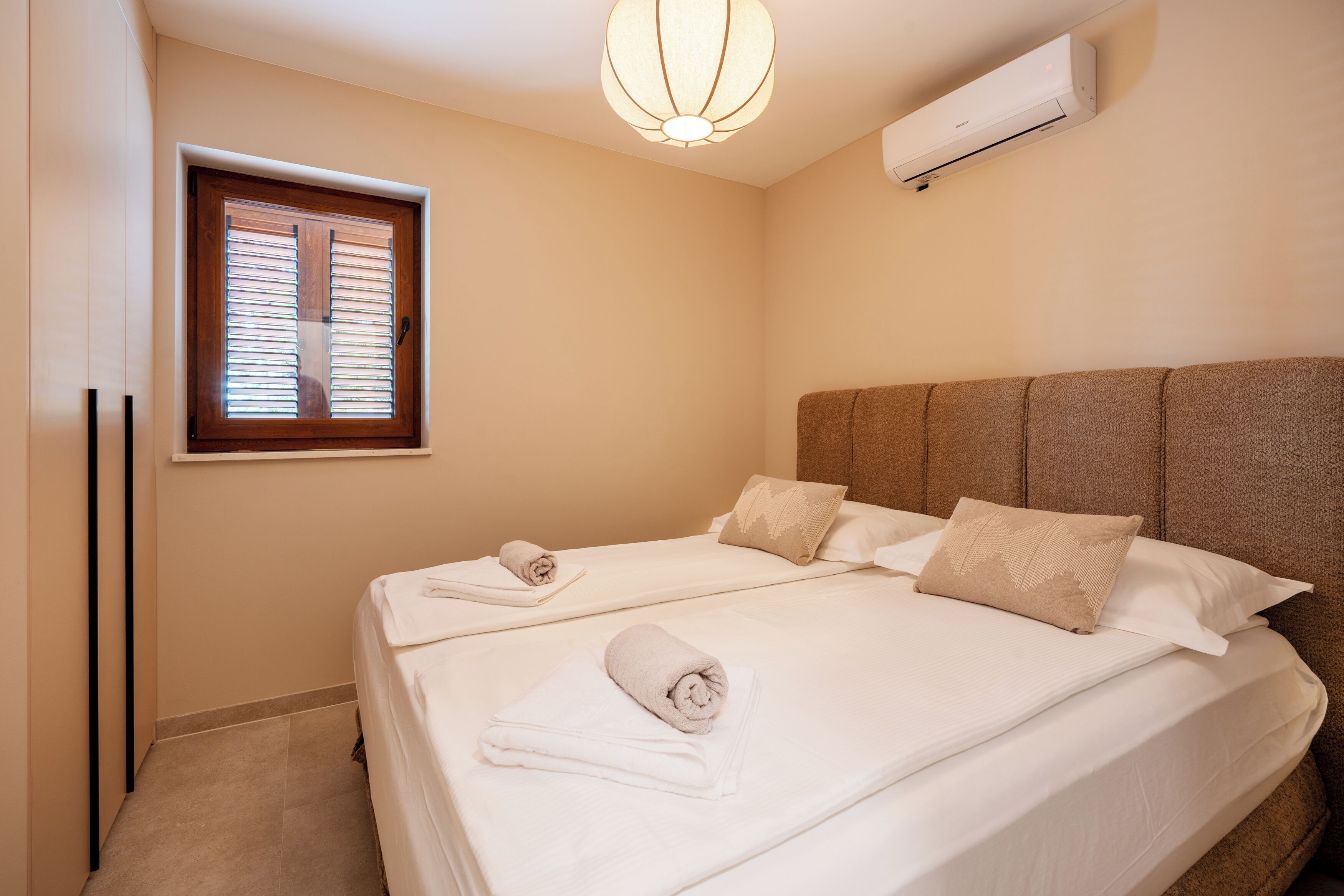 A bedroom with two beds, a window with blinds, a wall-mounted air conditioner, and a ceiling light fixture.